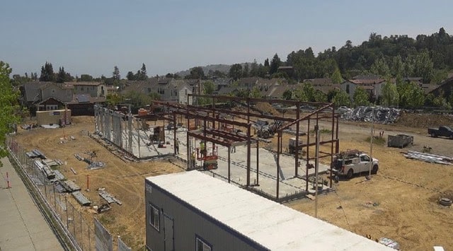 A construction site with a metal building frame being assembled, surrounded by dirt, construction materials, and vehicles; houses and trees are visible in the background.