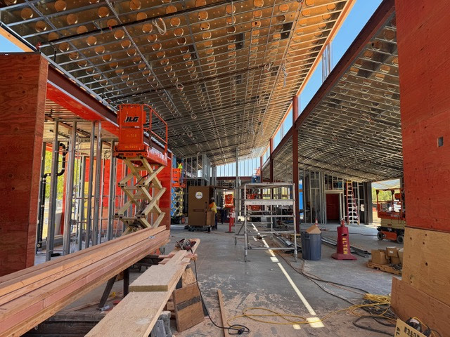 Interior view of a building under construction with exposed steel beams, plywood, scaffolding, construction equipment, and materials scattered on the ground. Open walls and ceiling reveal framework and unfinished structure.