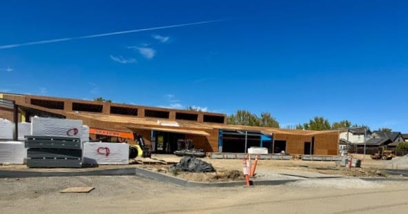 A single-story building under construction with wooden framework, construction materials stacked in the foreground, and a clear blue sky overhead. Some trees and houses are visible in the background.