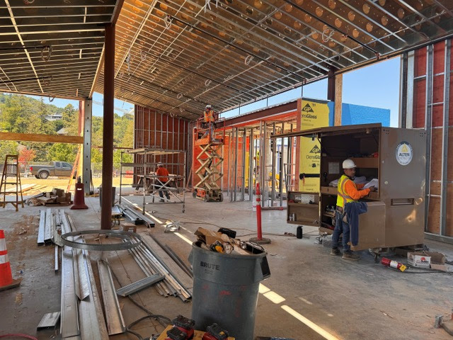 Construction site interior with exposed beams and framing. Two workers in safety gear are present—one on a scissor lift working near the ceiling, and another by a tool cabinet. Various building materials and tools are scattered around.