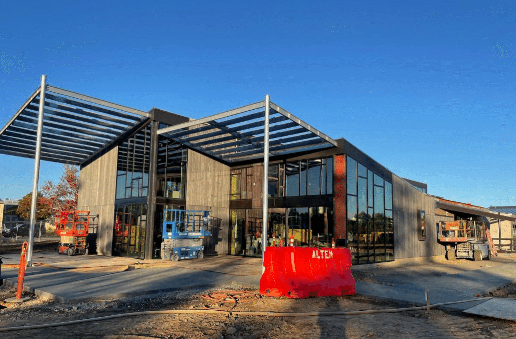A modern building under construction with large glass windows and metal frames, surrounded by construction equipment and a bright red barricade labeled "ALTER" in the foreground. The sky is clear and blue.