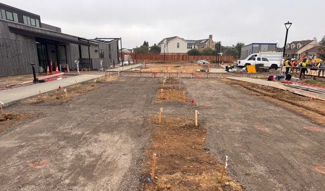 A construction site with dirt and gravel, wooden stakes marking the ground, workers in safety vests, a white truck, and nearby buildings. Equipment and traffic cones are visible on the left side.