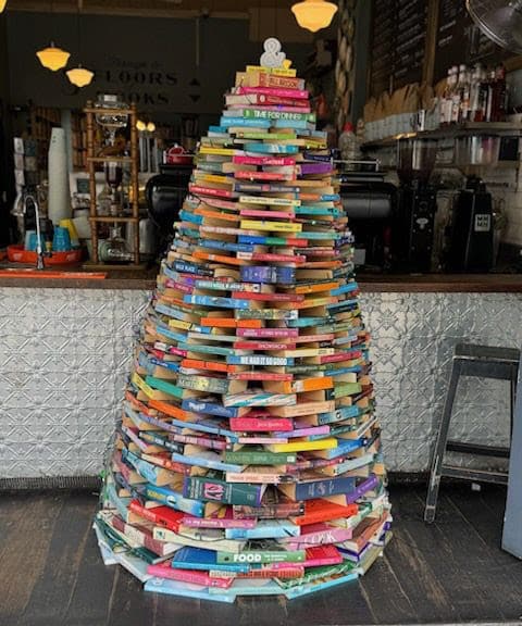 A Christmas tree made entirely of stacked, colorful books sits in front of a counter at a cozy café or bookstore. The books are arranged in circular layers, forming a tree shape.
