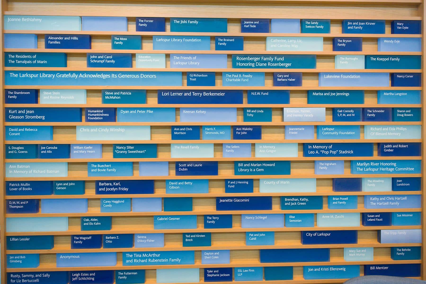 A wall display features numerous rectangular blue plaques of varying shades, each listing names of donors and families, arranged in a grid pattern against a wooden background at The Larkspur Library.