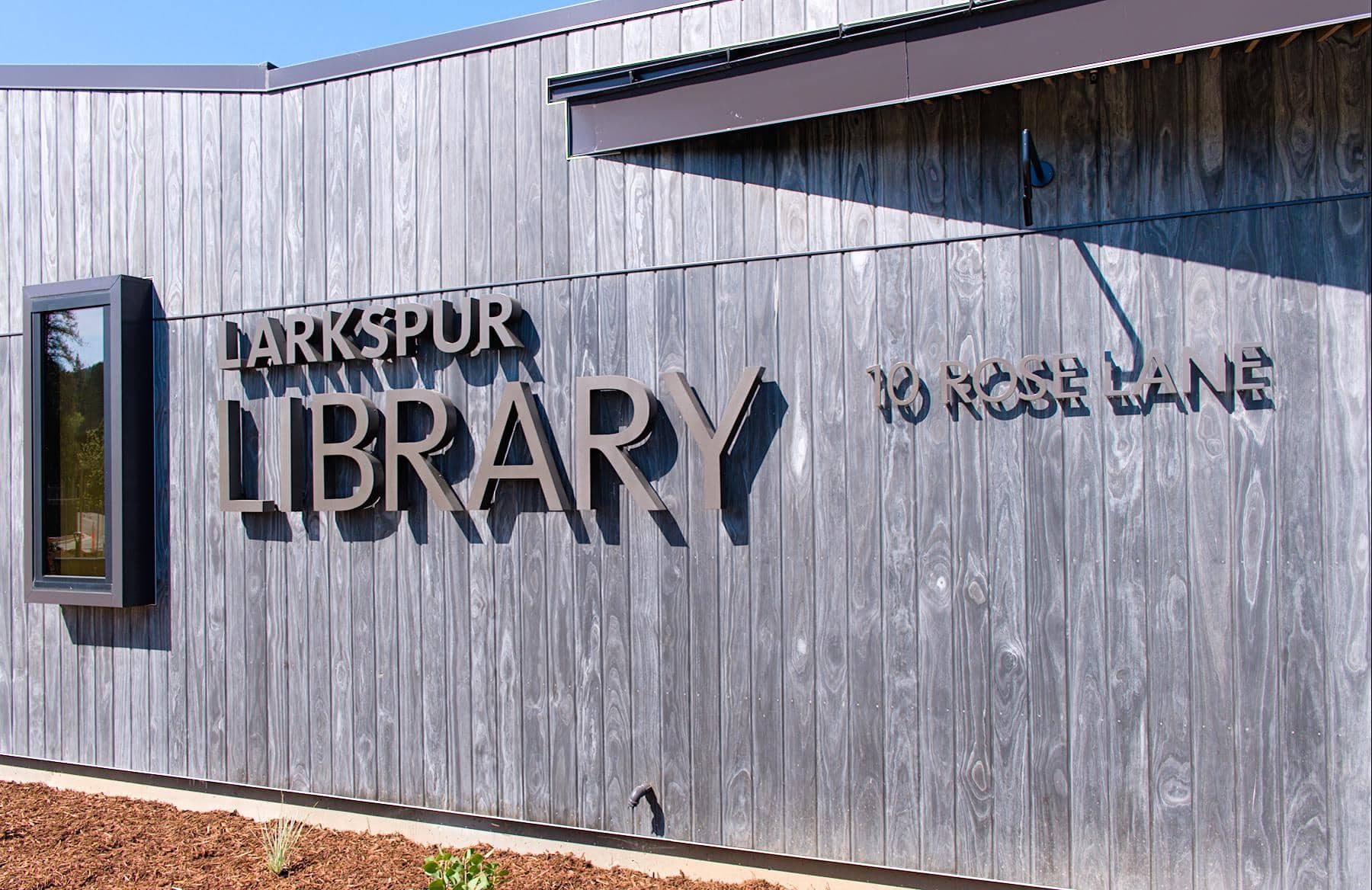 The exterior of our modern wooden building with large metallic letters spelling "Larkspur Library" and the address "10 Rose Lane" on the wall. A small rectangular window juts out to the left.