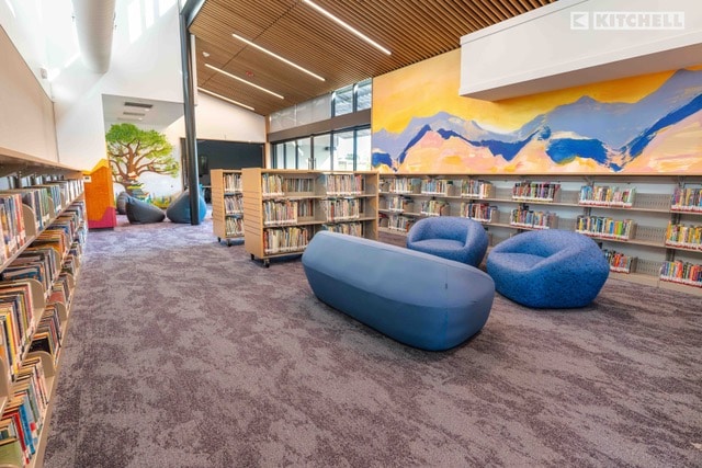 A modern library interior with bookshelves, soft blue seating, carpeted floors, large windows, and colorful wall murals of a tree and mountains. Natural light fills the space, creating a bright and inviting atmosphere.