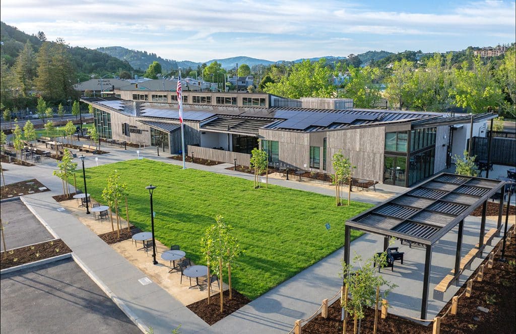 Drone image of the new modern library in Larkspur, with large windows and solar panels sits behind a green lawn with young trees, benches, and walking paths. Hills and trees are visible in the background, and the sky is bright and clear.