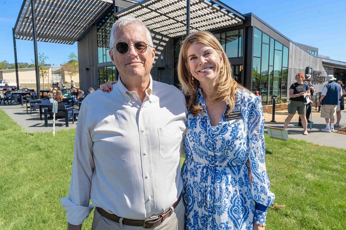 A man in sunglasses and a light shirt stands next to a smiling woman in a blue-and-white dress on grass outside a modern building, with people sitting and walking in the background on a sunny day.