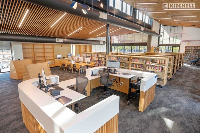 A modern library interior with large windows, wooden bookshelves filled with books, a curved central help desk with computers, and several workstations on a carpeted floor. Natural light fills the spacious room.