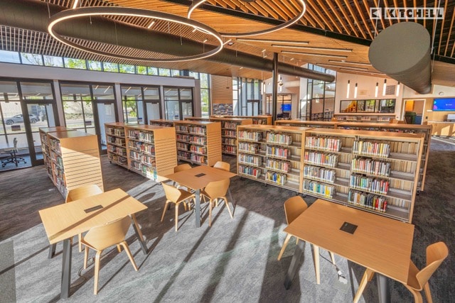 A modern library interior with wooden tables and chairs, bookshelves filled with books, large windows letting in natural light, and circular ceiling lights above.