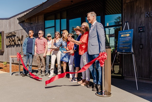 A group of people stand smiling in front of a building entrance, holding a large pair of scissors and ribbon for a ribbon-cutting ceremony. A sign and a cut red ribbon are visible in the foreground.