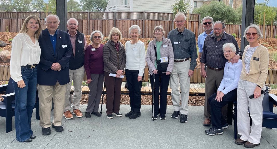 A group of thirteen older adults, some standing and some seated, pose together outdoors on a patio. They are smiling and casually dressed, with trees and a wooden fence in the background.