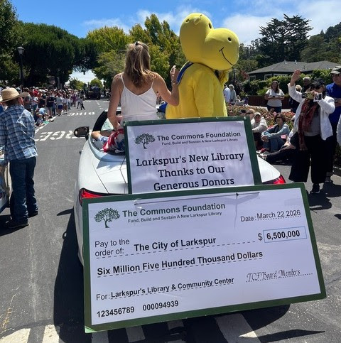 A woman and a yellow mascot sit on a car in a parade, holding giant checks for $6.5 million to fund Larkspur’s new library. People line the street, watching and cheering during the daytime event.