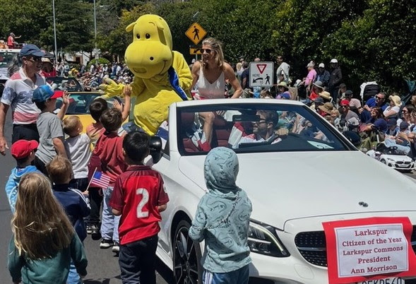 A parade scene with children gathered around a white convertible car. A person in a yellow dog costume and a woman ride in the car, as a crowd watches. A sign on the car names the Larkspur Citizen of the Year.
